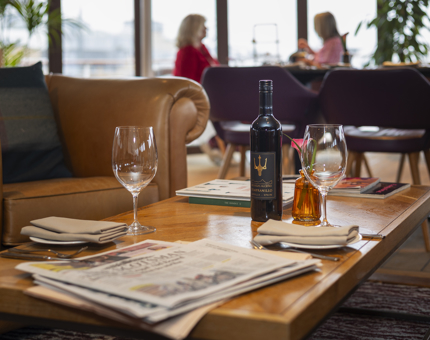 In The Lighthouse Restaurant, there are couches with a coffee table. On the table is a newspaper, two wine glasses and a bottle of wine. 