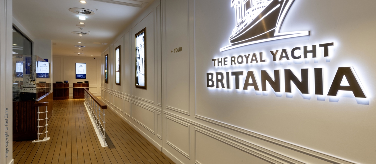 The entrance to the new Visitor Centre. An illuminated sign with the Britannia ship logo is on the wall. The flooring is replica decking and there are light box pictures further along the wall leading to the till points. 