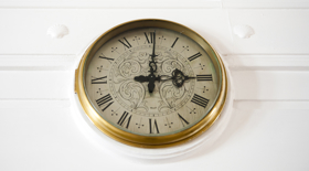 A gold‑rimmed wall clock with Roman numerals hangs on a white wall, its ornate black hands showing the time at three o’clock. A gold‑rimmed wall clock with Roman numerals hangs on a white wall, its ornate black hands showing the time at three o’clock.