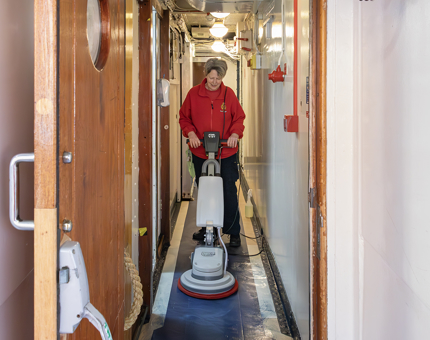 A woman using a floor buffing machine to polish the floors in the narrow corridor outside the Galley. 