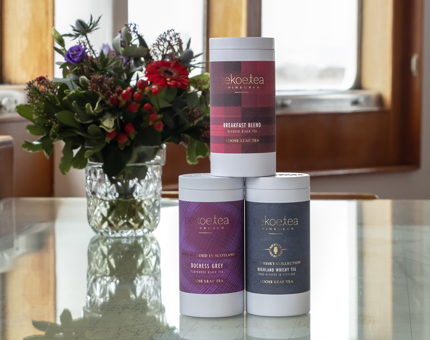 Three tins of tea with multi-coloured labels stacked in a pyramid. An arrangement of flowers is on the table behind. 