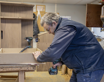 A man from the Maintenance team using a drill to assemble a wooden till point in the workshop. 