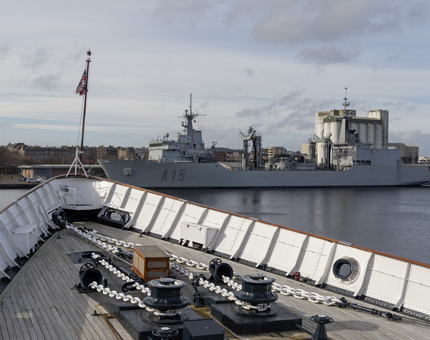 The bow of Britannia and a large grey naval ship in port in the background. 