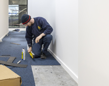 Another man from Facilities laying carpet tiles and using a spirit level to give a straight edge. 