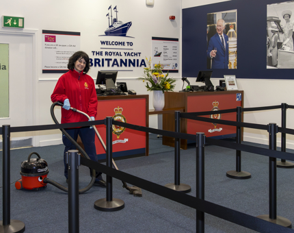 A Housekeeper is vacuuming inside the Ticket Office. There are two desks in the background and queue barriers. 