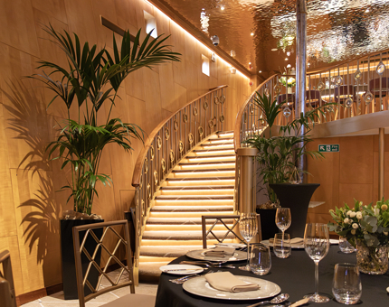 The sweeping staircase in the Ballroom at Fingal. A round table is set for dinner in the foreground and there are palm plants at the bottom of the stairs. 