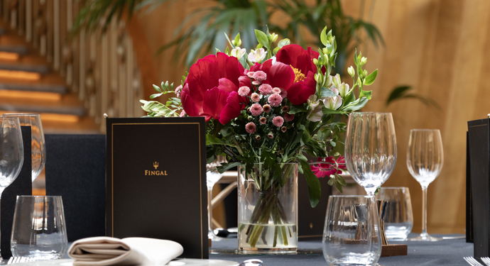 A table laid for dinner in Fingal's Ballroom. A vase of red and white flowers is on the table with menus and glasses. A sweeping staircase is in the background. 