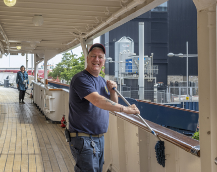 Man wearing navy blue t-shirt and trousers uses a duster to clean the side of the ship.