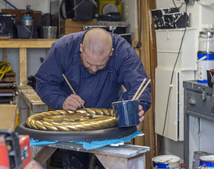 A man in a workshop holding a paintbrush and painting a sign. 