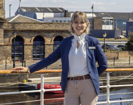 A woman standing with her hand on the railing of a ship's deck with water and a building behind her. 