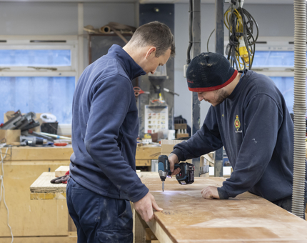 Two men in a workshop, one is holding a drill. 