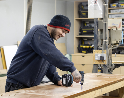 A man in a workshop drilling a table top. 
