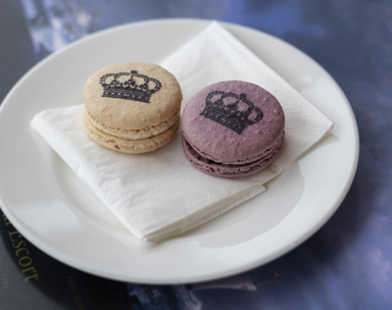 Two macarons on a plate, one purple and one orange with a picture of a crown on them. 