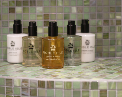 Five bottles of Noble Isle shower gel, shampoo and body lotion siting on a green tiled shelf. 