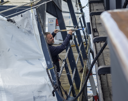 A man standing on a Paint Cat painting the side of the ship. 