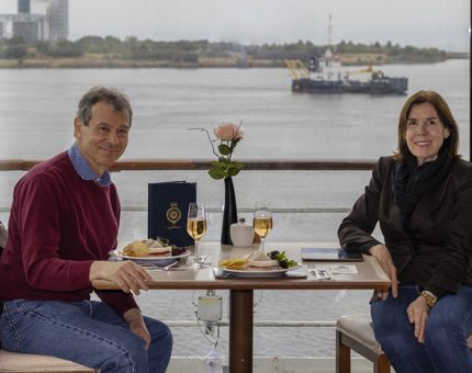 A man and a woman sitting at a table with plates of sandwiches, in front of a large window with views of water and boats. 