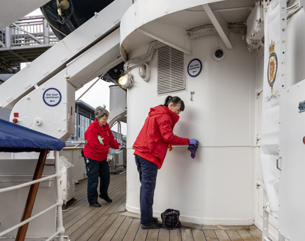 Two members of the Housekeeping team wearing red jackets applying polish to the brass handrail outside on a deck. 