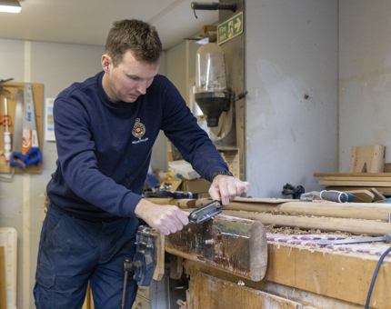 A Maintenance team member using a spokeshave to remove old varnish from a handrail.