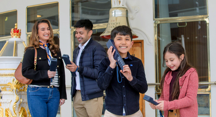 A family listening to an audio guide handset next to Britannia's Bell and binnacle.