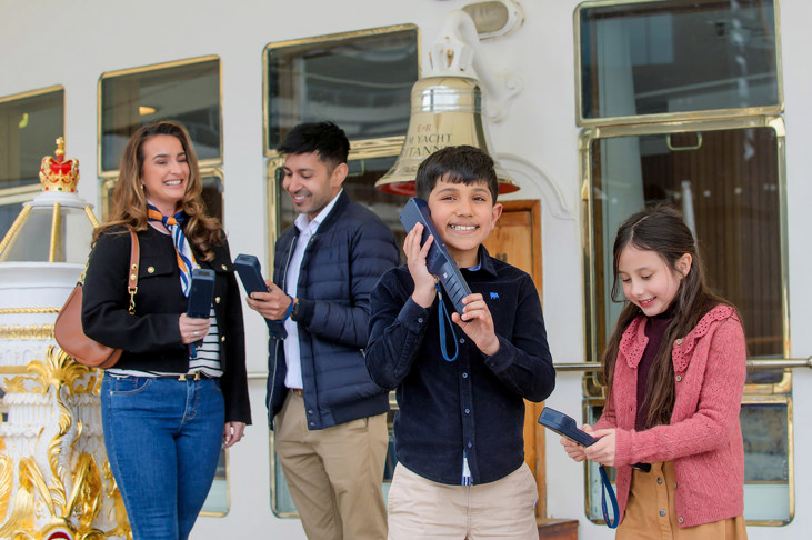 Four people, a woman, man, boy and girl stand on the Verandah Deck of Britannia, they are holding audio guide handsets.