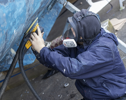 A man wearing a full face safety mask holding a sander. He is sanding the blue paint off of the side of a boat. 