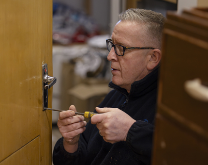 A lock being repaired on a door. A man is holding a screw driver. 