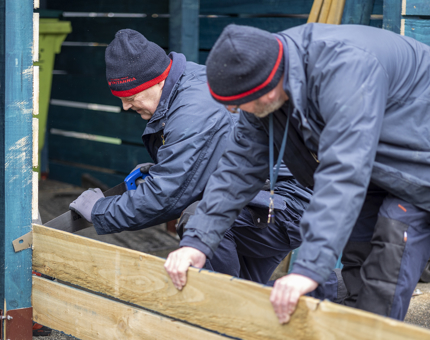 Two of the Facilities Officers making storage. One is holding a plank of wood, the other is using a saw to cut. 