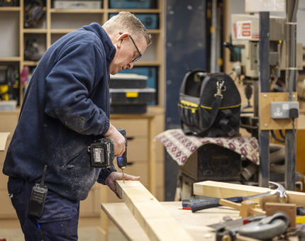 A maintenance team member holding a drill and drilling into a piece of wood. 