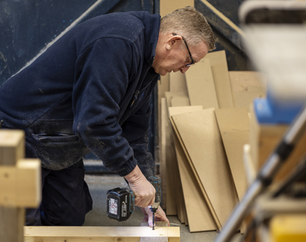 A closer look at a Maintenance team member drilling into a piece of wood with a drill. 