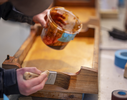 A close up oh hands holding a pot of varnish and a paintbrush to varninsh wooden steps for the Fast Motor Launch. 