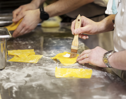 A close up of two pairs of hands preparing ravioli. The person in the foreground is holding a pastry brush. 