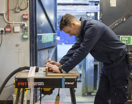 A Maintenance man is at a workbench cutting pieces of wood. 