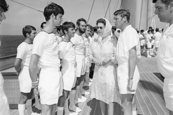 A black and white photo of a woman in a dress speaking to yachtsman on the deck of a ship 
