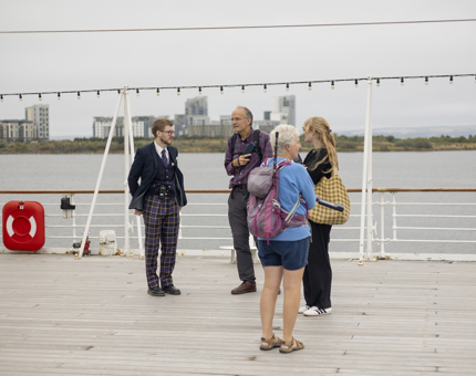 A Visitor Assistant is standing on Britannia's Verandah Deck talking to 3 visitors. The Port of Leith is in the background. 