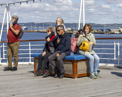 Visitors are sitting on a bench on the Verandah Deck in Leith. They are listening to audio guide handsets. 