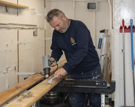Varnish being removed from wooden handrails with a heat gun and scraper being held by a Maintenance man.