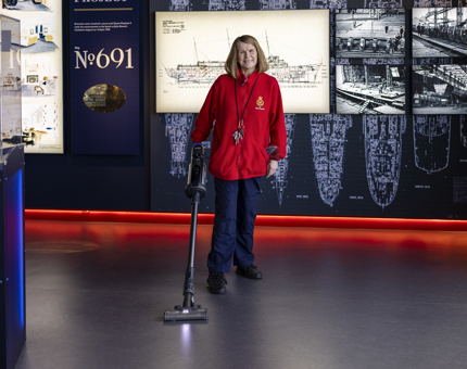 A Houskeeper is hoovering the floor of Britannia's Visitor Centre. There are images of ship drawings behind her on the wall. 