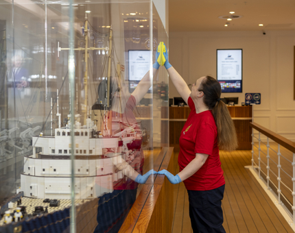 A Housekeeper is polishing the glass of a case containing a Lego model of Britannia. 
