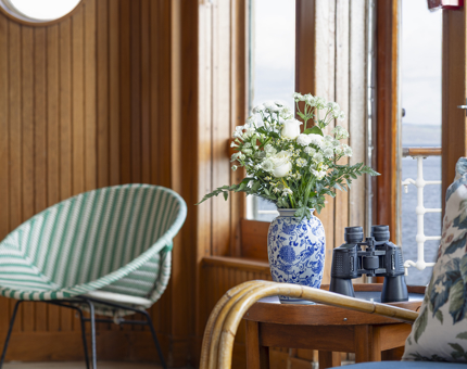 A detailed look at a section of the Sun Lounge, there is a round green and white striped chair and a table with a vase of flowers and binoculars on it. 