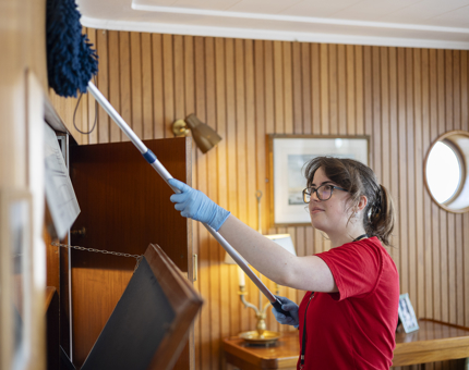 A Housekeeper is dusting in the Sun Lounge with a long blue duster.