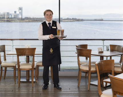 A hospitality server standing in front of the windows in Britannia's Tearoom holding a tray of drinks. 