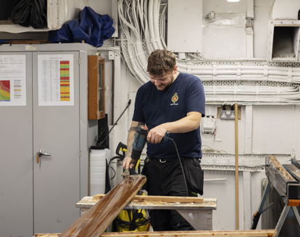 A maintenance team member is in the workshop removing varnish from a handrail using a heat gun. 