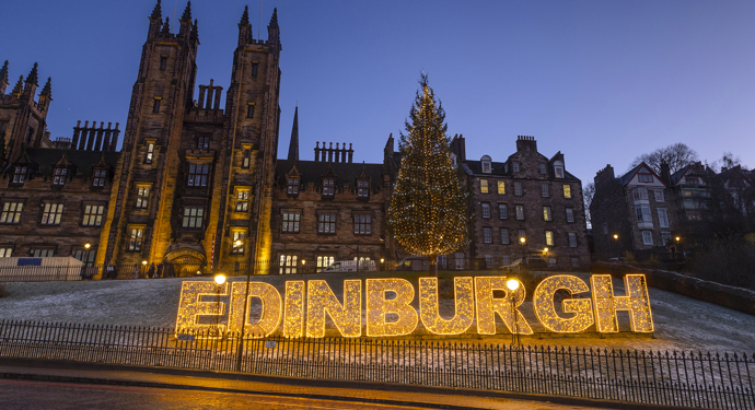 The word Edinburgh made up of lights. There are historic buildings in the background. 