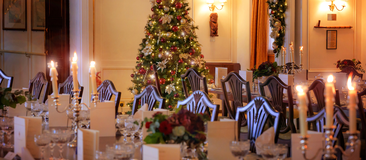 Dining room table set for an event with candles lit and a Christmas tree with gold and red decorations in the background