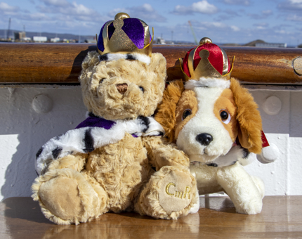 A cuddly souvenir teddy bear wearing a crown and cloak and a cuddly King Charles spaniel dog wearing a crown from the Britannia Gift Shop.