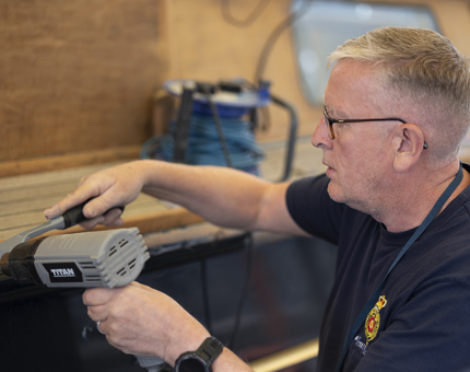 A man holding a heat gun, scraping varnish off of the Royal Barge. 