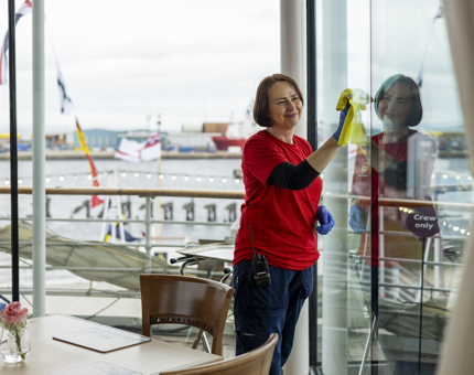 A Housekeeper in the Royal Deck Tearoom cleaning the windows. There is a view of the Port of Leith behind her. 