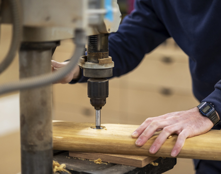 A member of Maintenance is using machinery to drill a hole through a wooden handrail. 