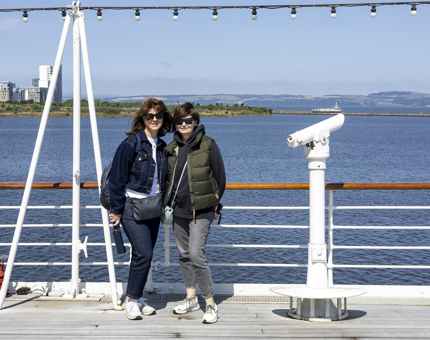 Two Visitors stop for a photo on the Verandah Deck with the Port of Leith behind them. 