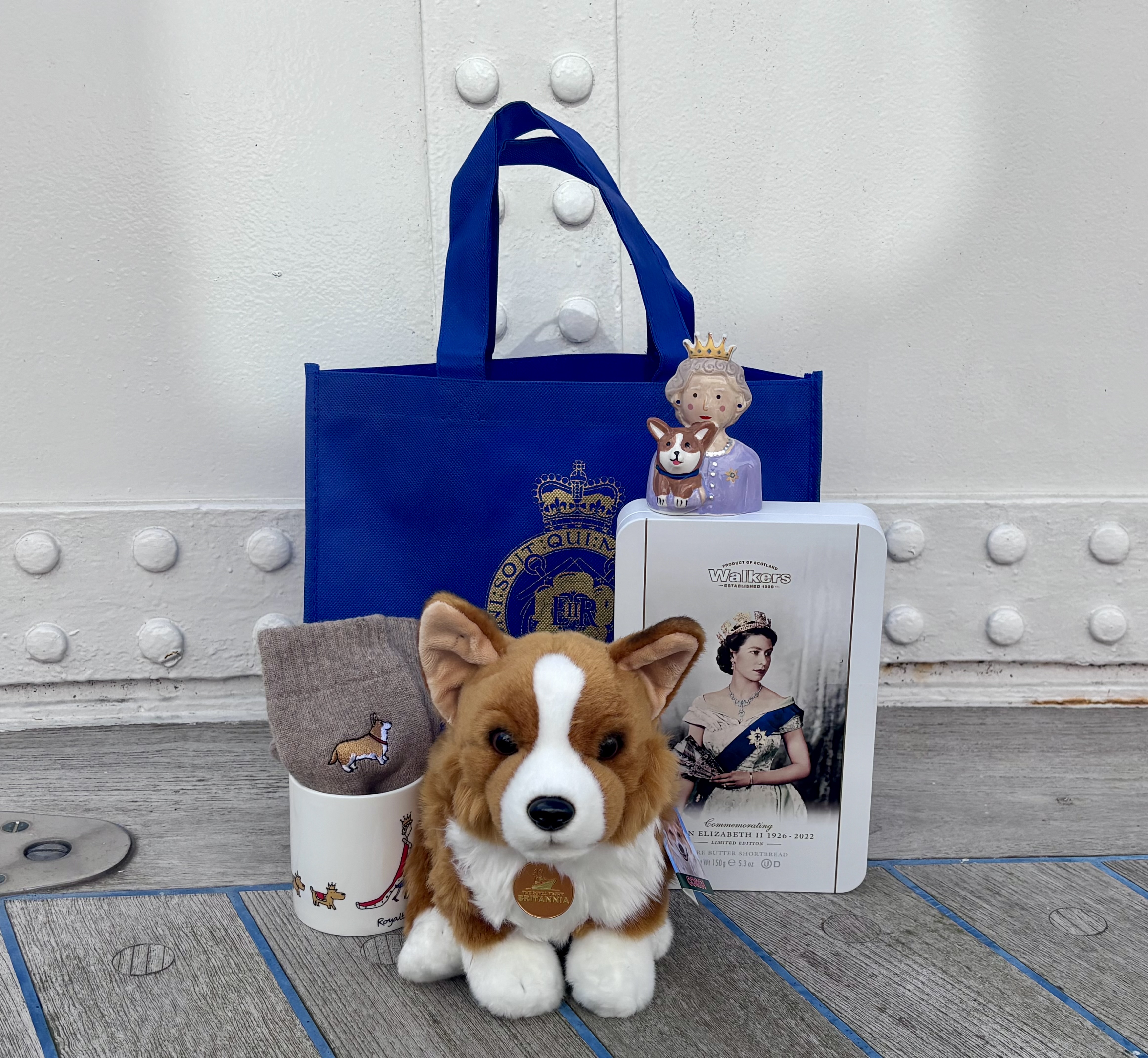 A neatly arranged display of Royal Yacht Britannia souvenirs on a wooden surface, including a blue tote bag with a gold crest, a plush corgi, a corgi‑patterned mug, grey corgi socks, a figurine of Queen Elizabeth II holding a corgi, and a booklet featuring her portrait.